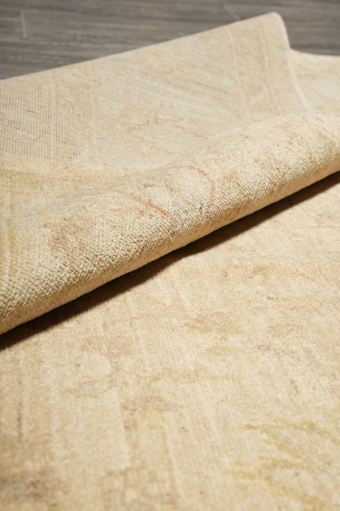 A close-up of a light beige textured rug with a rolled corner showing its woven pile and subtle tonal pattern, placed on a gray wood floor.