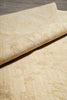 Load image into Gallery viewer, A close-up of a light beige textured rug with a rolled corner showing its woven pile and subtle tonal pattern, placed on a gray wood floor.