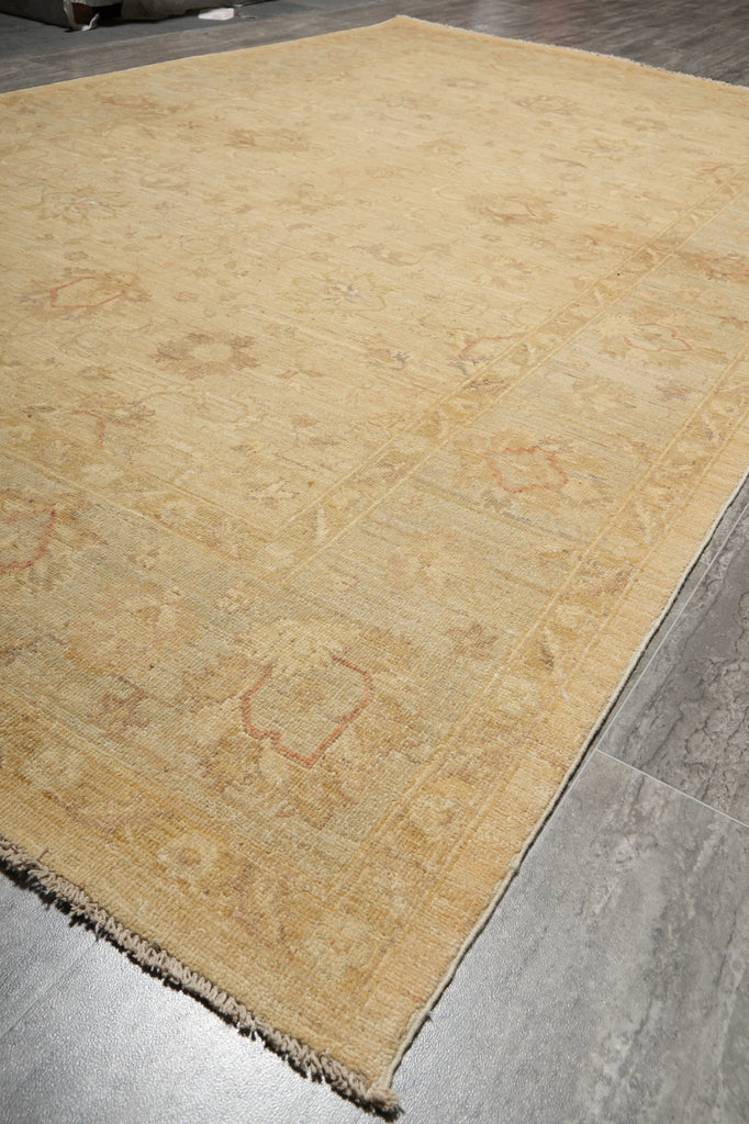 A large beige area rug with a faded traditional floral pattern and fringed edges, shown laid out on a gray tiled floor.