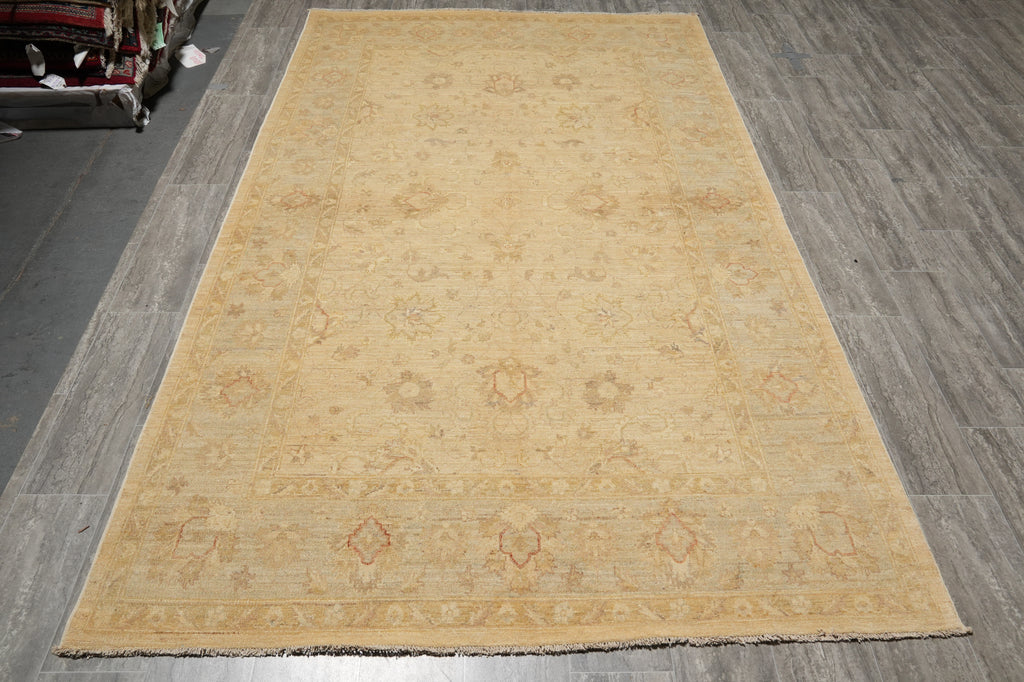 A large rectangular area rug in a warm sandy beige color with a faded traditional floral pattern, laid out on a gray wood-look floor.