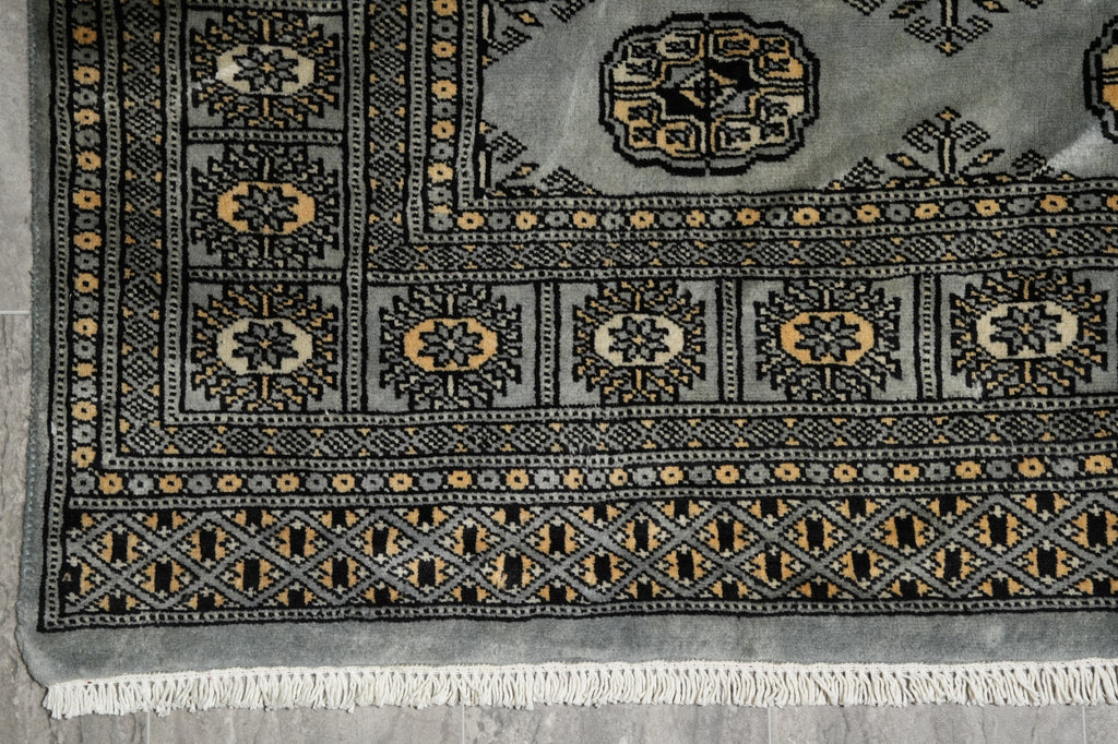 A close-up of a patterned area rug showing its corner, detailed medallion motifs, geometric borders, and white fringe tassels on a gray background.