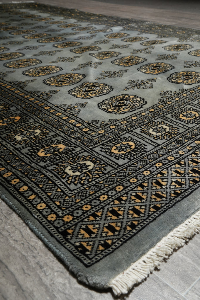 A close-up of a patterned area rug with a gray base, black geometric borders, and gold and beige floral motifs, showing a fringed edge.