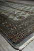 Load image into Gallery viewer, A close-up of a patterned area rug with a gray base, black geometric borders, and gold and beige floral motifs, showing a fringed edge.
