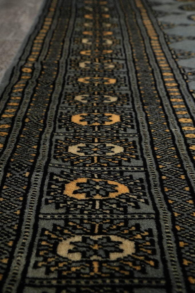 Close-up of a patterned textile or rug showing a dark charcoal base with repeating geometric medallion motifs in beige and muted gold.