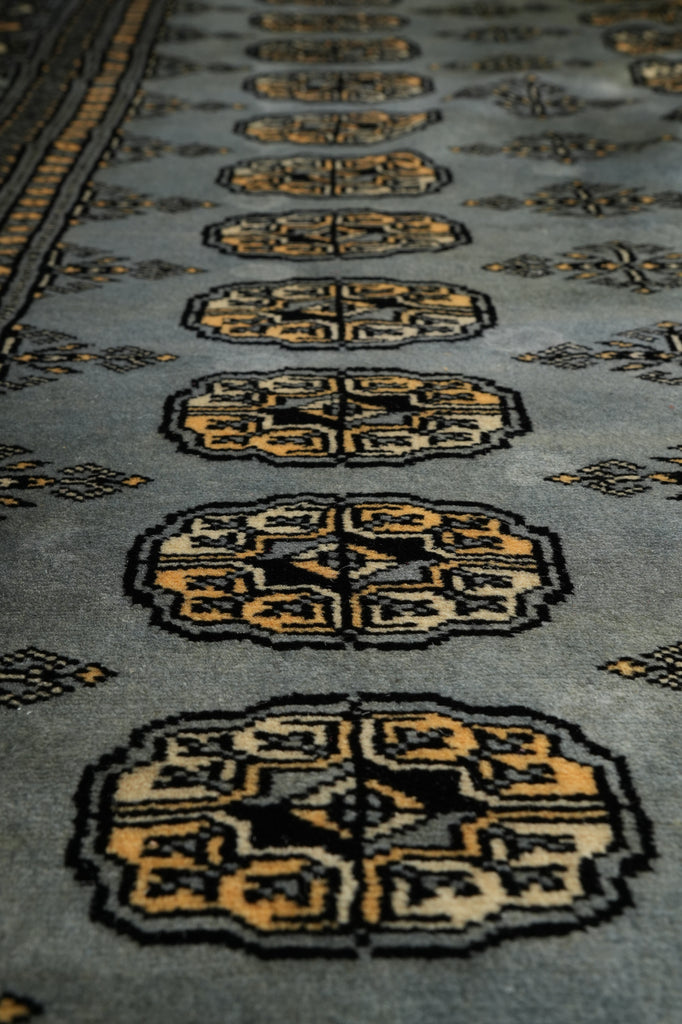 Close-up of a woven rug with repeating geometric medallion motifs in beige, black, and muted gold on a gray-blue background.