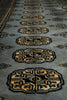 Load image into Gallery viewer, Close-up of a woven rug with repeating geometric medallion motifs in beige, black, and muted gold on a gray-blue background.