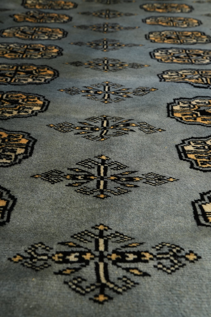 A close-up of a woven rug or textile with a muted blue-gray base and repeating geometric motifs in black and warm beige/gold.