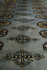 Load image into Gallery viewer, A close-up of a woven rug or textile with a muted blue-gray base and repeating geometric motifs in black and warm beige/gold.