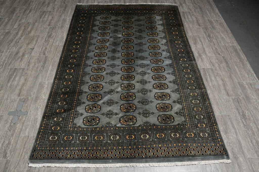 A rectangular area rug with a dark gray and black base, featuring a central field of repeating gold and brown medallions and ornate borders, laid out on a gray wood-look floor.