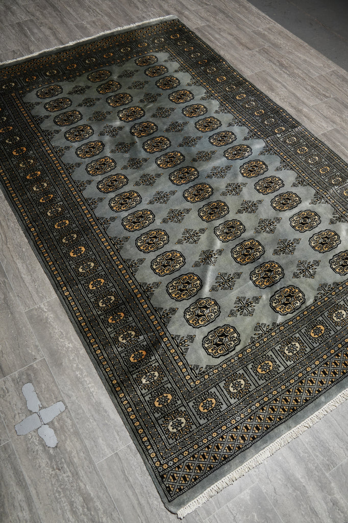 A rectangular area rug with a gray-blue field, black border, and repeating gold and brown medallion motifs, shown laid out on a gray wood-look floor.