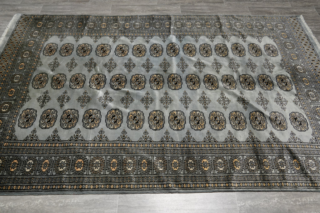 A large rectangular area rug with a light gray field, detailed medallion motifs, and a dark border, laid flat on a gray tiled floor.