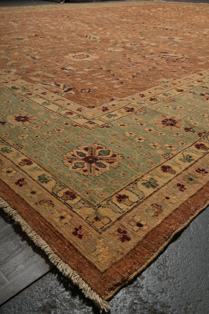 A traditional oriental area rug with a warm brown field, a muted sage green border, and cream fringe, shown laid out on a dark gray floor.