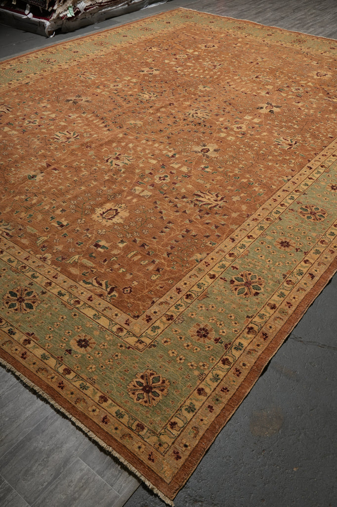 A large rectangular area rug with an ornate traditional pattern, featuring a warm rust-orange center field and a muted sage green border, laid out on a dark gray floor.