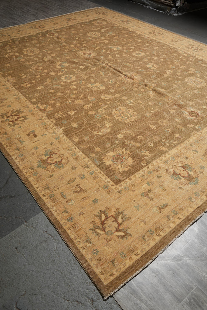 A large traditional area rug with a warm tan and beige base, featuring a central medallion pattern and a decorative border in muted brown, green, and cream tones, laid out on a gray stone floor.