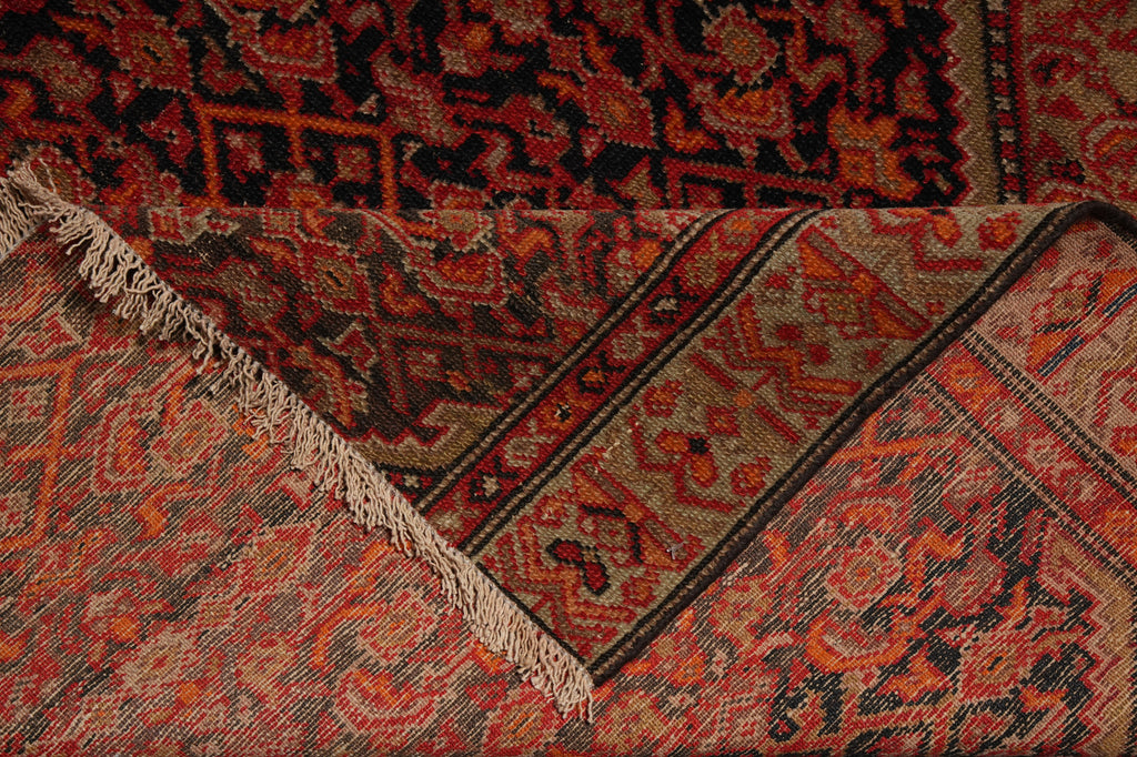 A close-up of a patterned rug with a folded corner showing its beige and rust-red woven design, dark border, and cream fringe.