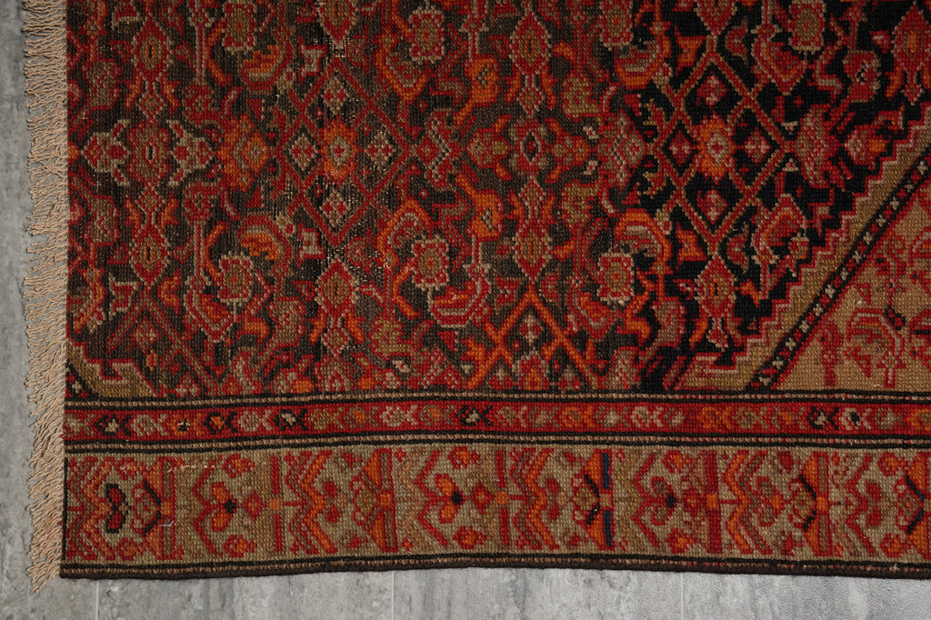 A close-up of a patterned rug showing a dark red and black central field with a beige and red geometric border, finished with a fringed edge on the left.