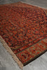 Load image into Gallery viewer, A close-up of a patterned area rug with a fringed edge, shown laid on a gray floor. The rug features a complex traditional design in red, orange, brown, and beige tones.