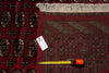Load image into Gallery viewer, A close-up of a patterned area rug in deep red and black, showing its woven geometric design, cream fringe, and a yellow tape measure laid across the corner with a white tag attached.