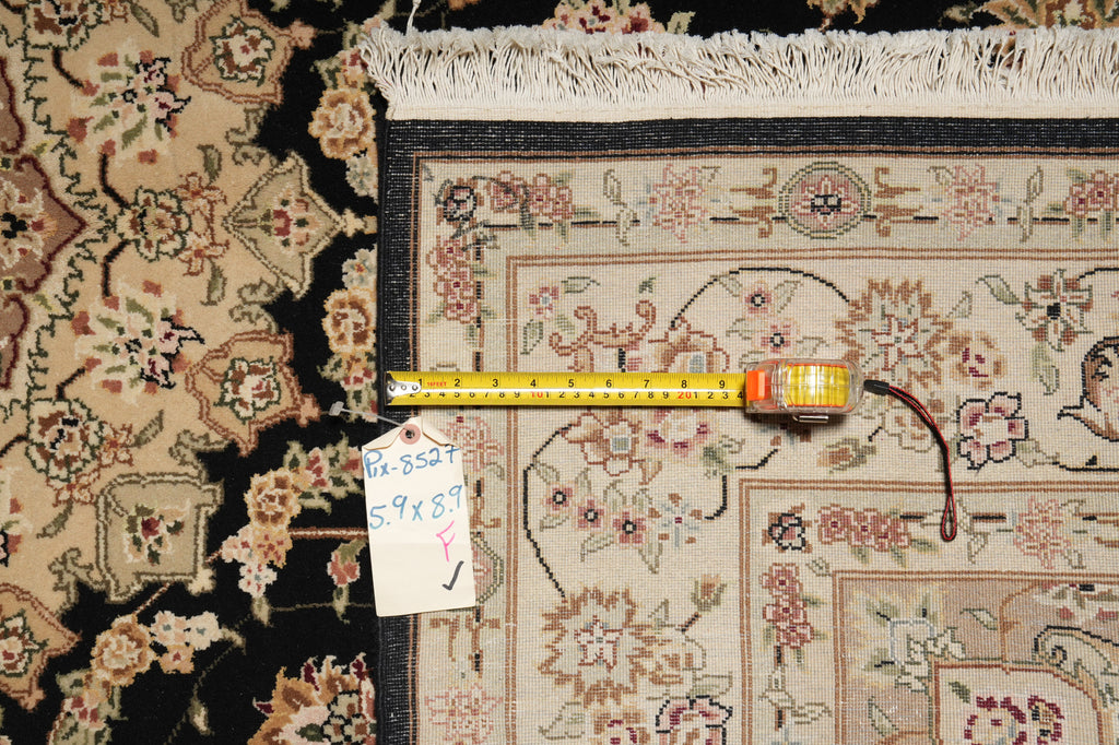 A close-up of a patterned area rug corner with a tape measure laid across it and a handwritten size tag attached. The rug has a cream and beige base with floral motifs in pink, green, and brown, plus a dark border.