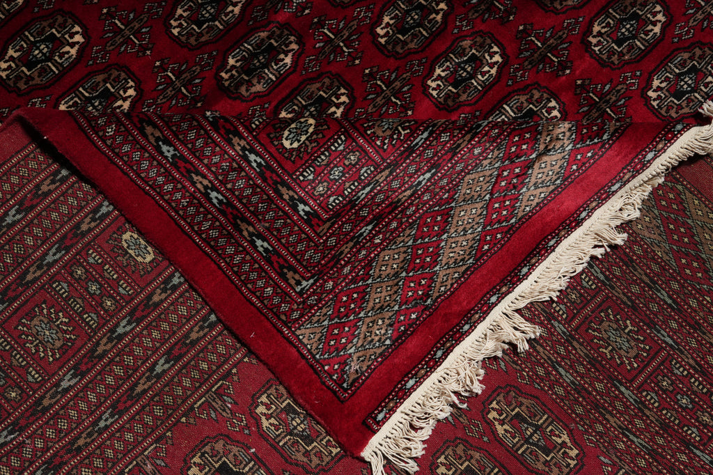 A close-up of a traditional patterned rug with a deep red field, geometric borders, and off-white fringe.