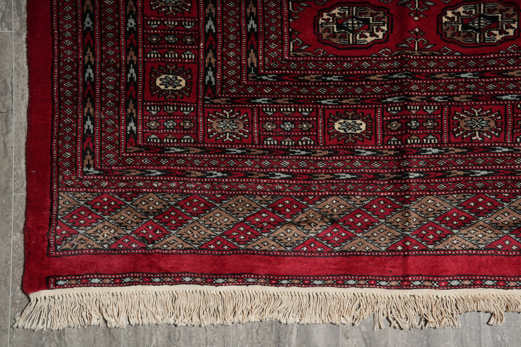 A close-up of a traditional patterned rug corner with a deep red field, geometric borders, and cream fringe along the bottom edge.