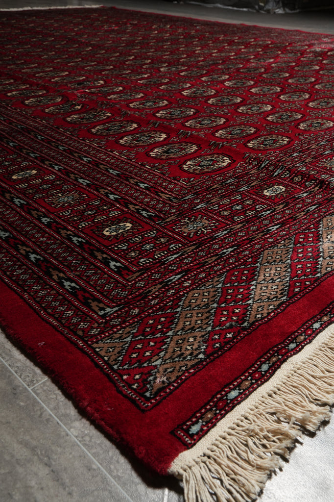 A close-up of a traditional patterned rug with a deep red field, cream fringe, and beige, blue, and black geometric motifs.