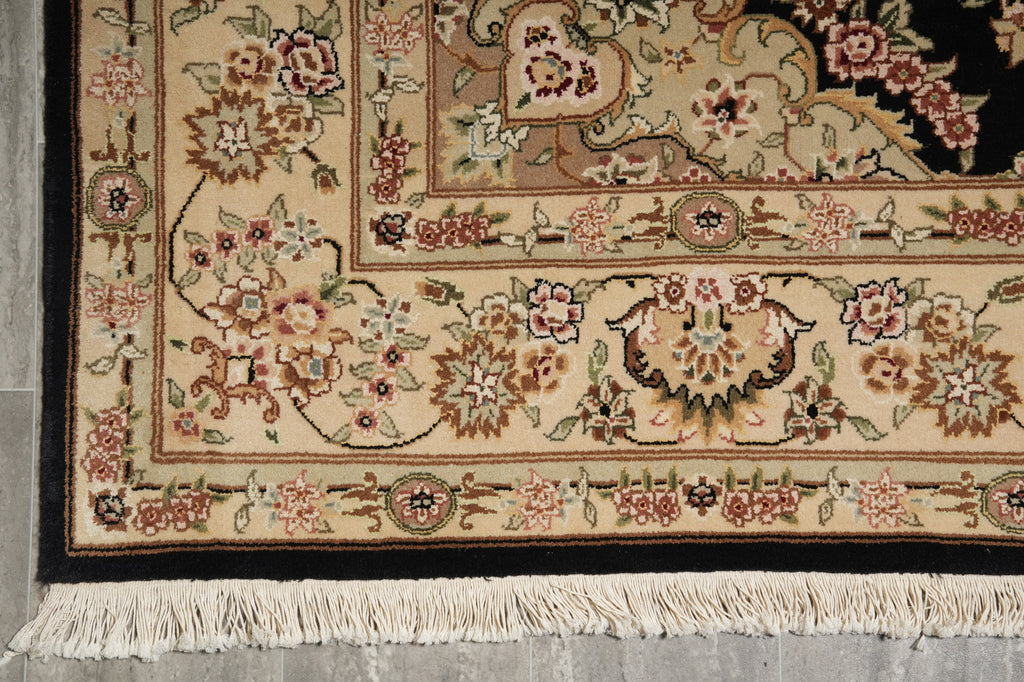 Close-up of an ornate area rug corner showing a cream and beige floral pattern with pink, green, and brown details, a dark border, and white fringe on a gray floor.