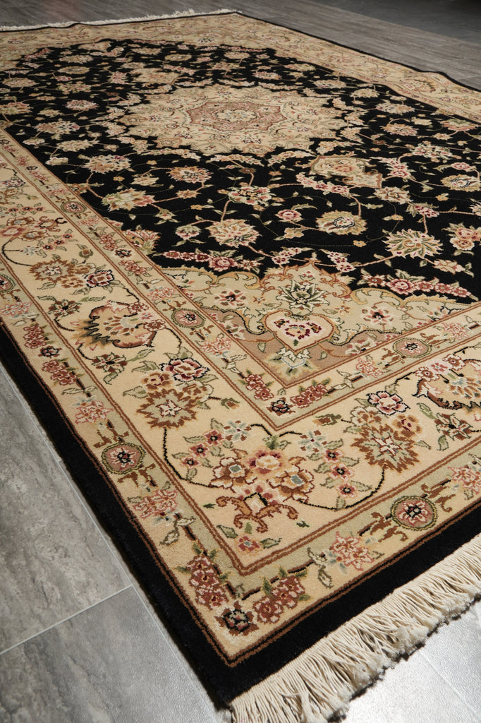 A traditional oriental-style area rug with a black field and beige border, featuring intricate floral patterns in muted pink, green, and gold tones, laid out on a gray tiled floor.