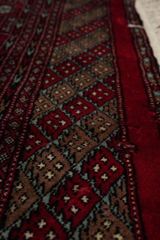 A close-up of a patterned rug with geometric diamond motifs in deep red, brown, black, and cream, showing the woven texture and fringe along the bottom edge.