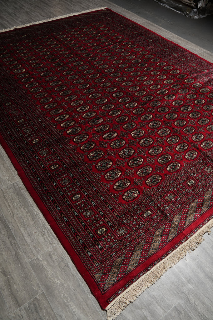 A traditional Persian-style area rug with a deep red field, intricate geometric and floral motifs in beige and black, and cream fringe on the ends, shown laid out on a gray tiled floor.