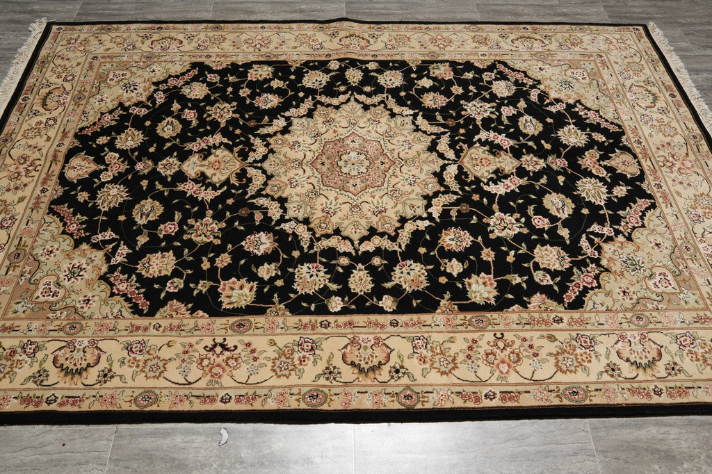 A large traditional oriental area rug with an intricate floral pattern, featuring a black central field, beige borders, and multicolored floral details, laid out on a gray tiled floor.