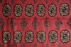 Load image into Gallery viewer, A close-up of a red patterned rug or textile featuring repeating geometric medallion motifs in black, beige, and muted gold tones.