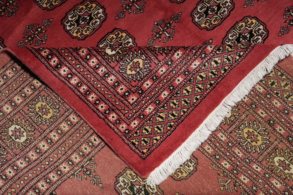A close-up of a patterned rug with a deep red field, intricate geometric and floral motifs in black, beige, and gold, and a white fringe along one corner.