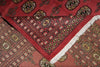 Load image into Gallery viewer, A close-up of a patterned rug with a deep red field, intricate geometric and floral motifs in black, beige, and gold, and a white fringe along one corner.