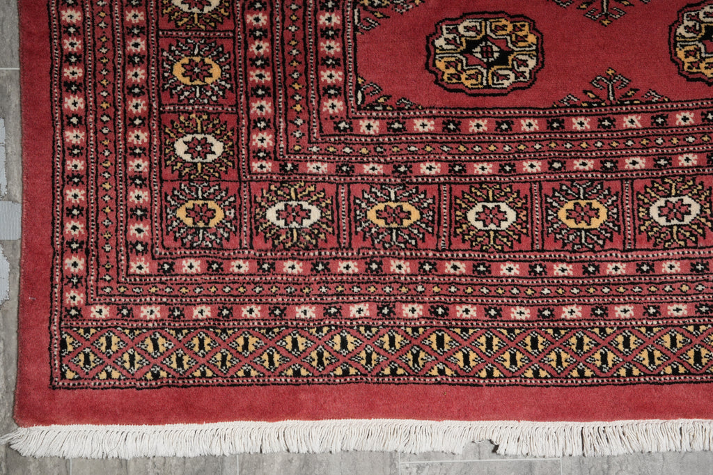 A close-up of a red patterned rug with white fringe, featuring geometric borders and floral motifs in black, beige, and yellow.