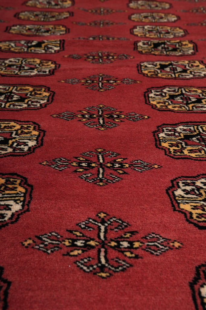 A close-up view of a red patterned rug with repeating geometric and floral motifs in black, beige, and muted yellow.