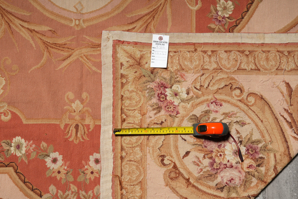 A close-up of a vintage-style floral tapestry rug with a dusty rose and beige background, featuring a yellow tape measure and a small white tag placed on it for scale.