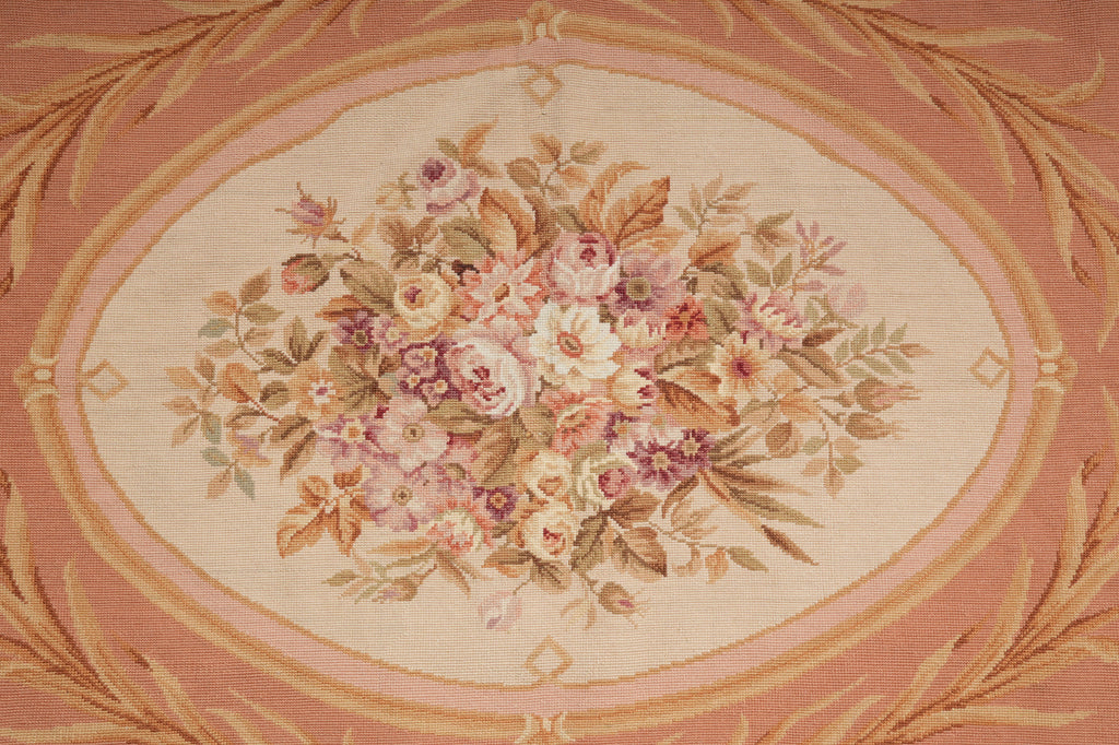 A close-up of a woven floral textile or rug showing an oval bouquet motif in soft pink, cream, beige, and muted green tones, surrounded by a peach border with decorative leafy detailing.