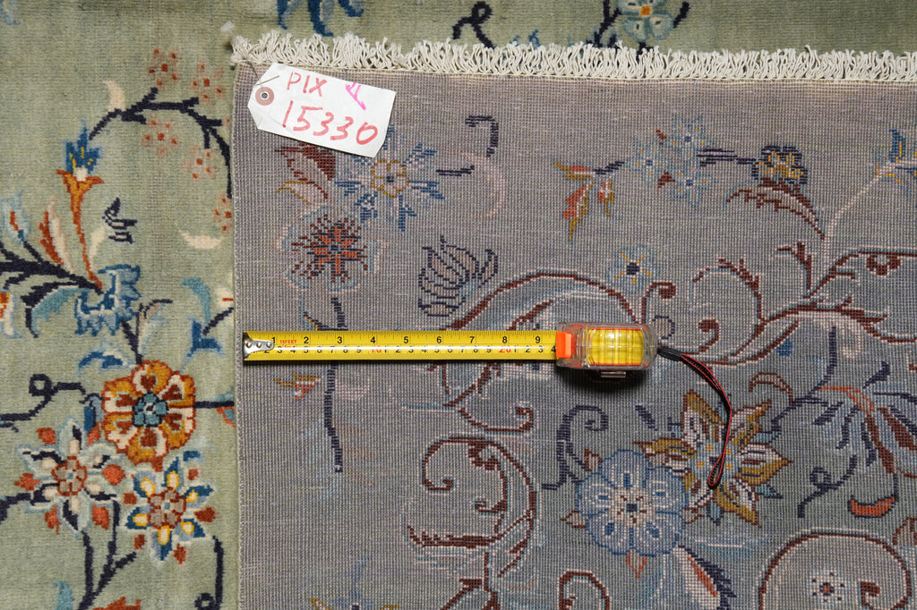 A close-up of a patterned area rug with a muted blue-gray base and floral motifs in orange, blue, and cream, shown with a yellow tape measure and a small inventory tag.