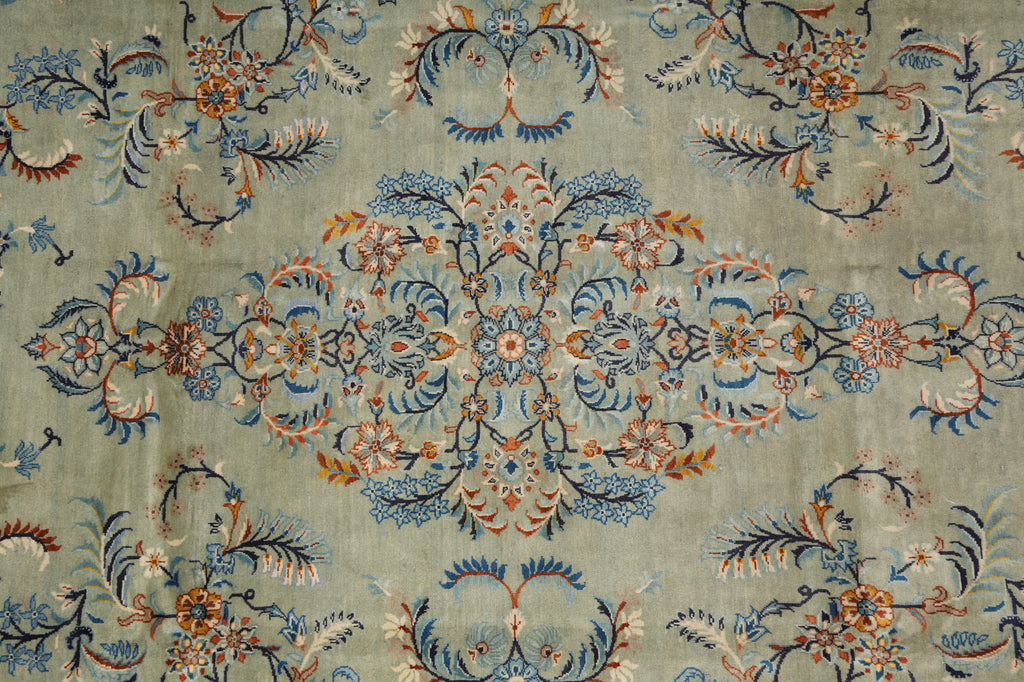 A close-up of an ornate floral patterned textile or rug with a muted sage green background and detailed motifs in blue, rust orange, cream, and dark navy.