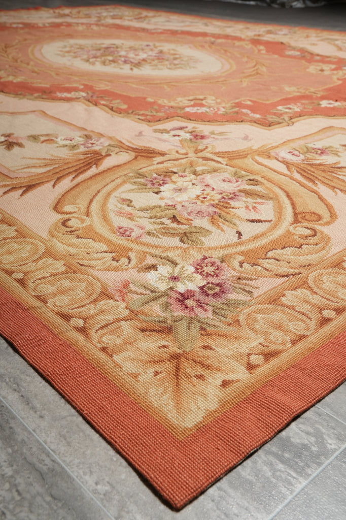 A close-up of an ornate area rug with a warm peach and beige floral pattern, featuring a solid rust-orange border, laid on a gray tiled floor.