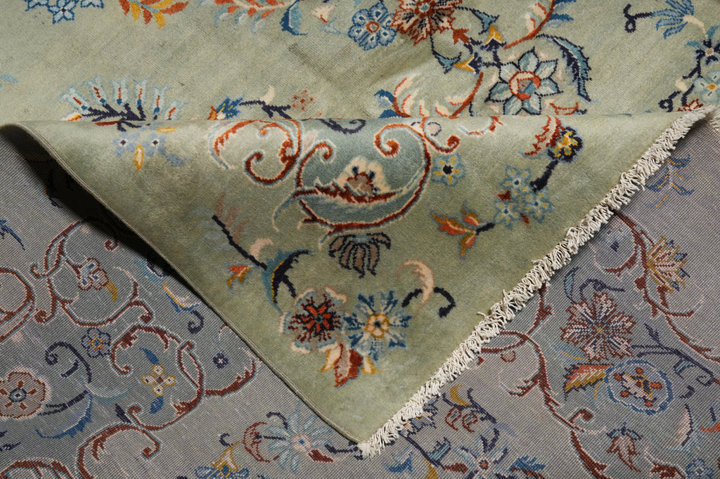 A close-up of a folded area rug showing its intricate floral and scrollwork pattern. The top layer is a pale sage green with multicolored motifs in blue, rust red, and orange, finished with white fringe.