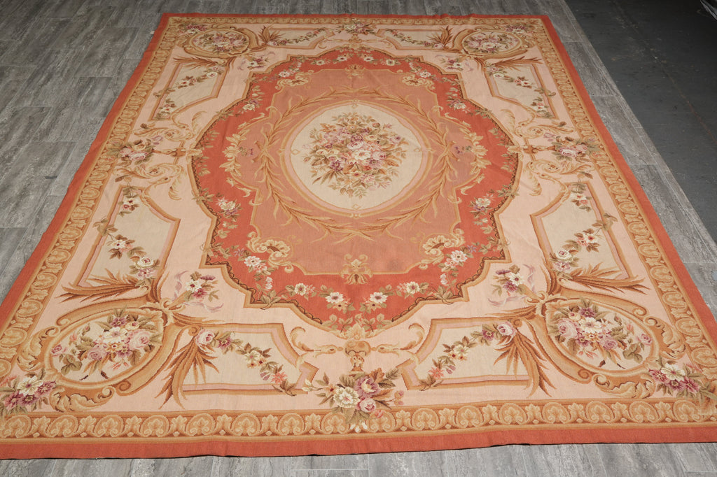 A large rectangular area rug with a classic French Empire or Louis XV style medallion pattern, laid out on a gray wood-plank floor. The rug features a warm peach and orange border, a central cream medallion with floral details, and ornate scrollwork in beige and gold tones.