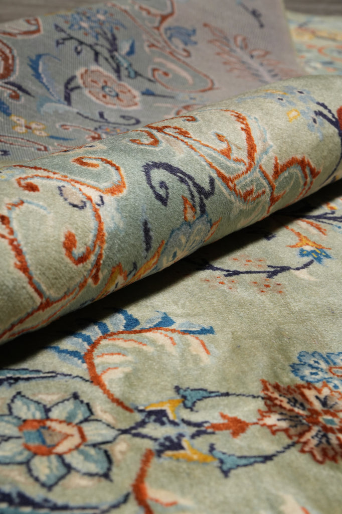 A close-up of a folded area rug showing its detailed pattern and pile texture. The rug features a muted sage green base with intricate floral and scroll motifs in blue, rust orange, and cream.