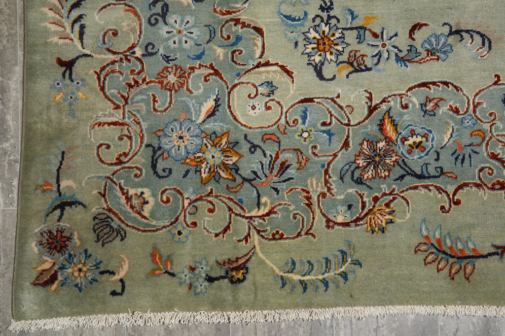 A close-up of an ornate floral rug with a sage green background, featuring intricate vine and flower patterns in blue, rust red, and gold, finished with white fringe.