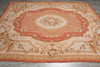 Load image into Gallery viewer, A large traditional French tapestry rug with an ornate central floral medallion, featuring a warm palette of terracotta red, beige, and gold on a gray tiled floor.
