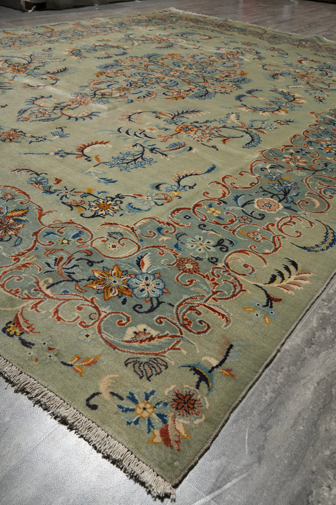 A traditional oriental area rug with a sage green base and intricate floral patterns in blue, orange, beige, and white, shown laid out on a gray concrete floor.