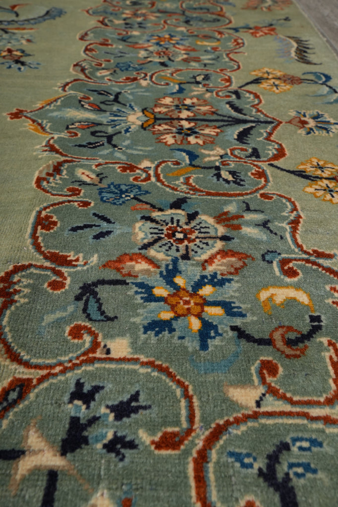 A close-up view of an ornate area rug with a traditional floral and scroll pattern on a muted sage green background, featuring accents of blue, rust red, orange, and cream.