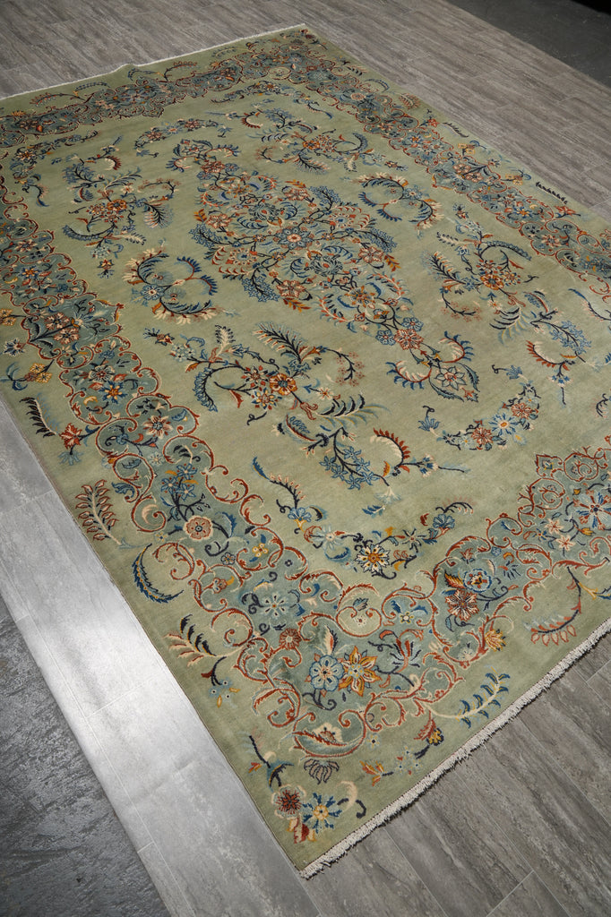 An ornate traditional area rug with a sage green base and intricate floral motifs in blue, orange, and beige, shown on a gray wood-look floor.