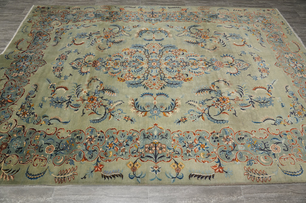 An ornate traditional area rug with a central medallion and floral motifs, featuring a muted sage green background with blue, rust orange, and beige details, laid out on a gray wood floor.